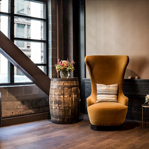 A stylish seating area with two mustard-yellow wing chairs, a black quilted chair, a barrel with flowers, and large windows ends the sentence.