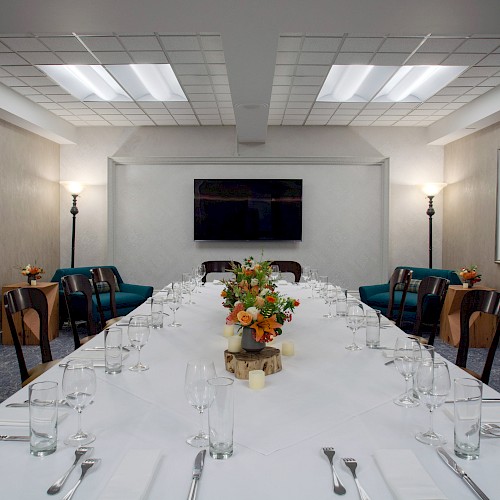 A long conference table set for dining, with flowers in the center, surrounded by chairs in a room with a TV and framed artwork on the wall.