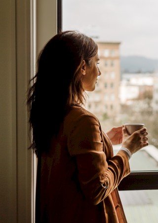 A person stands by a window holding a cup, looking at an urban view with several buildings visible in the background.