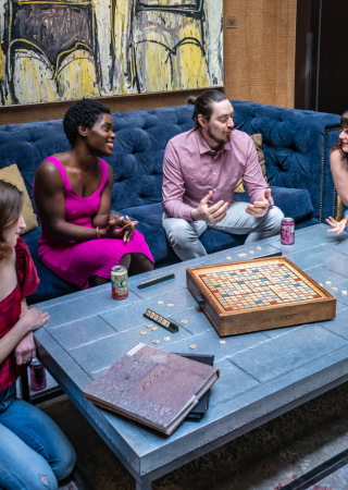 Four people are sitting and talking around a coffee table with a Scrabble game, books, and drinks in a cozy room.