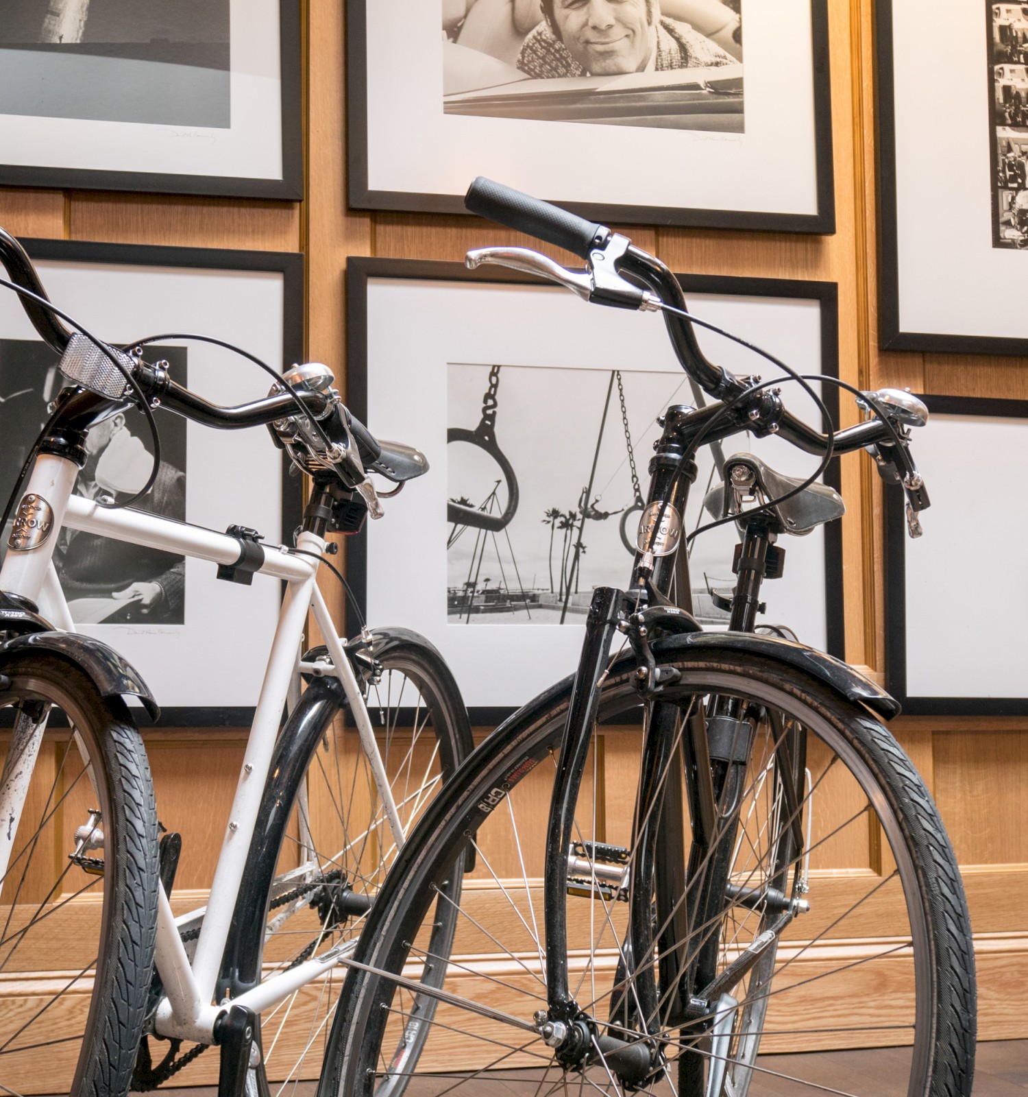 Two bicycles are parked indoors against a wall decorated with framed black and white photographs and artwork.