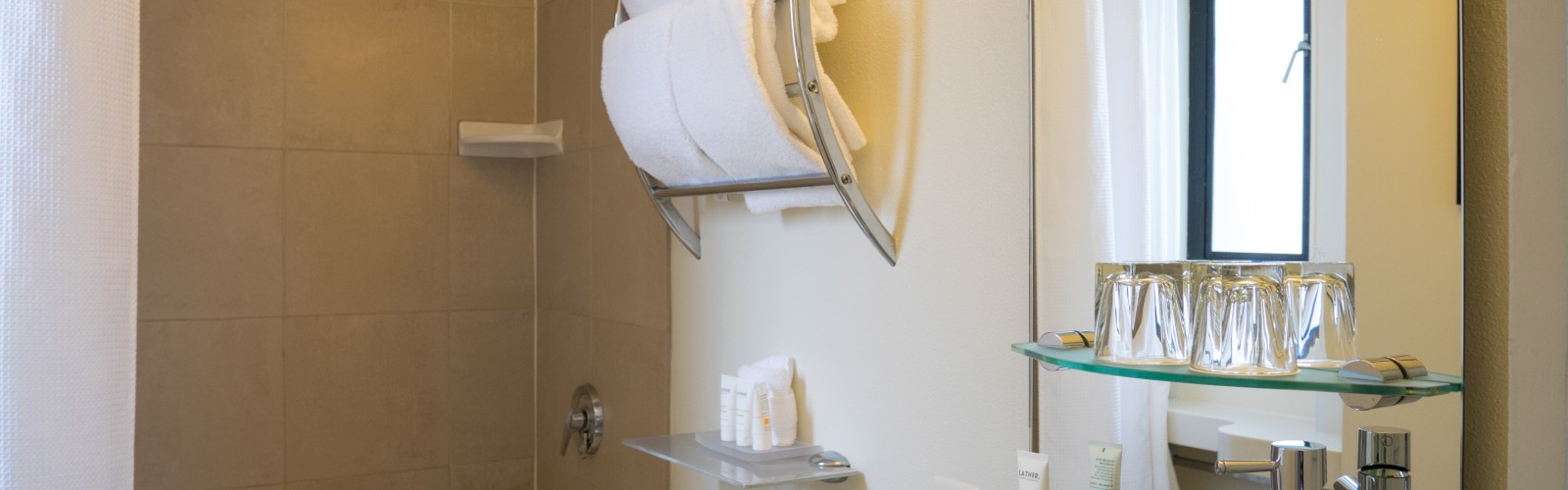 A modern bathroom with a bathtub, shower, towel rack, sink, mirror, toiletries, and glass shelf with cups.