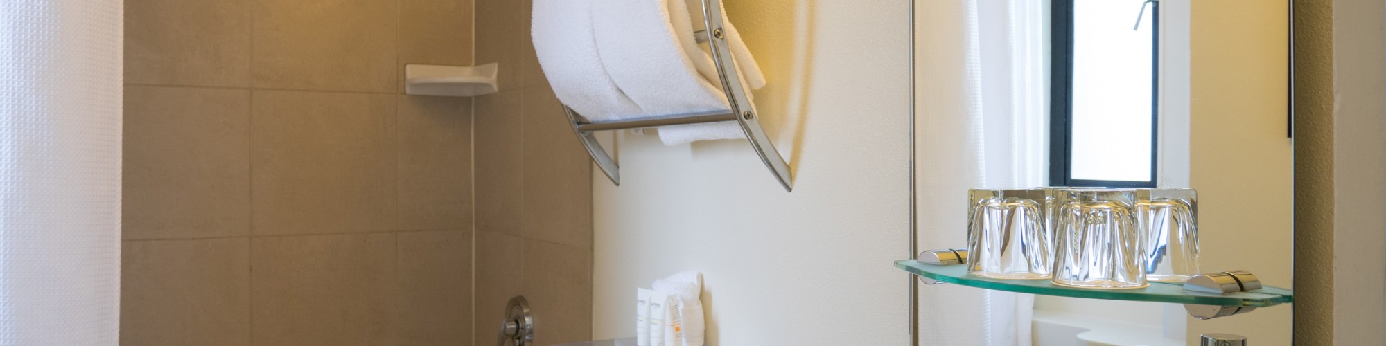 A modern bathroom with a bathtub, shower, towel rack, sink, mirror, toiletries, and glass shelf with cups.