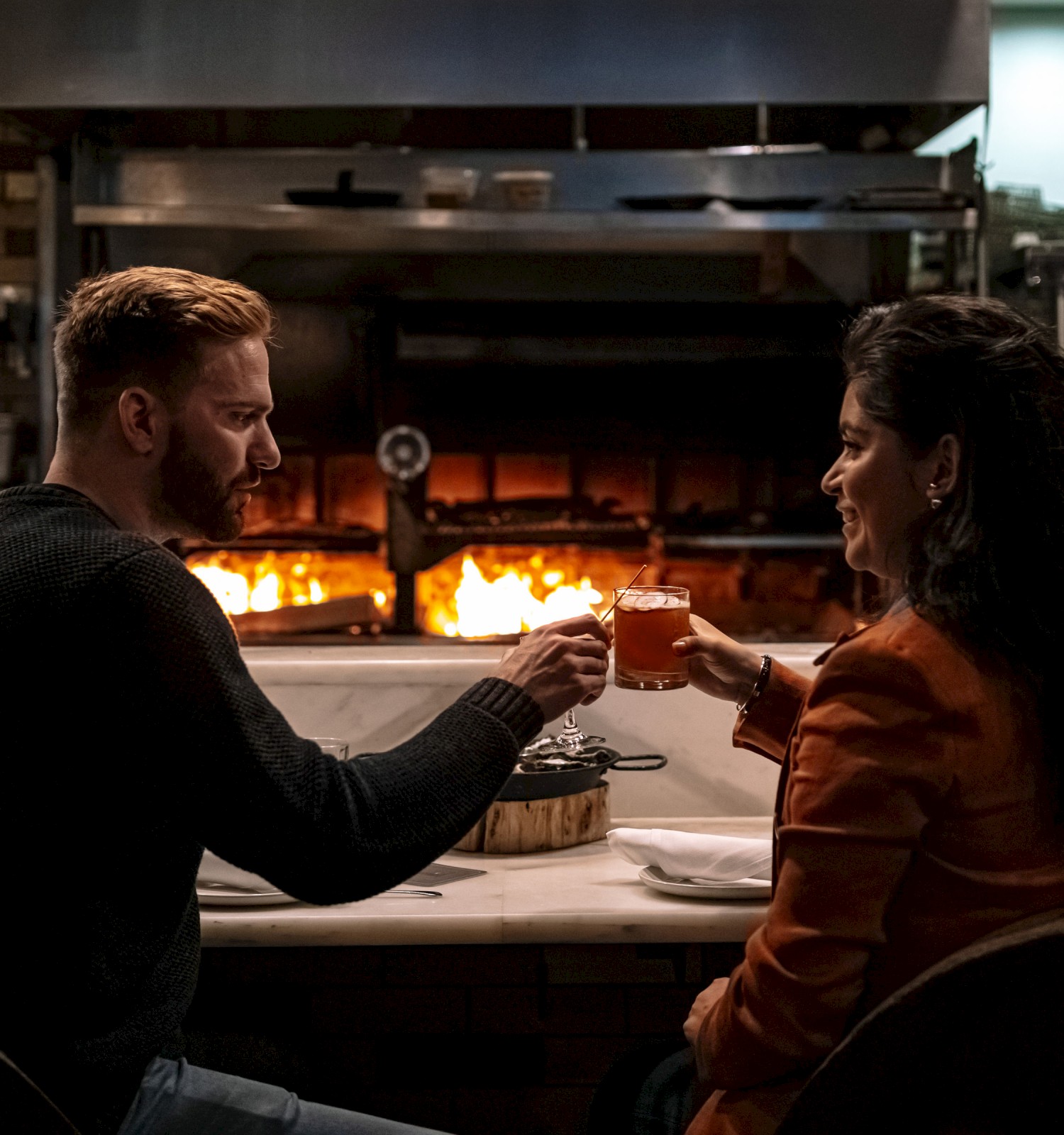 A couple is sitting in a cozy restaurant, toasting with drinks in front of a warm fireplace, creating a romantic atmosphere.