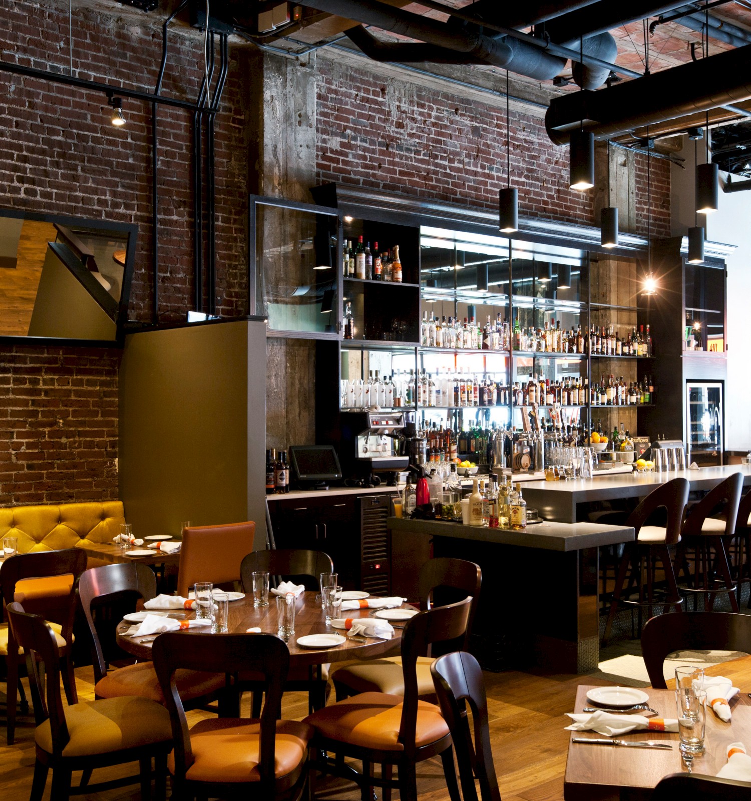 The image shows a modern restaurant interior with brick walls, a bar, wooden tables, and chairs, creating a cozy dining atmosphere.