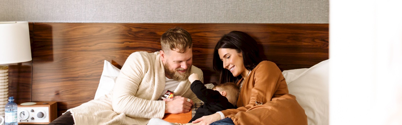 A family is joyfully sitting together on a bed, laughing and cuddling with their child in a cozy room setting.