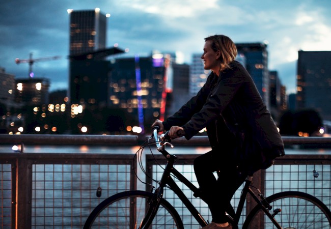 A person rides a bicycle on a bridge, with a city skyline and river in the background during sunrise or sunset.