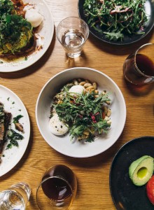 A variety of dishes including salads, eggs, avocado, and drinks are arranged on a wooden table.