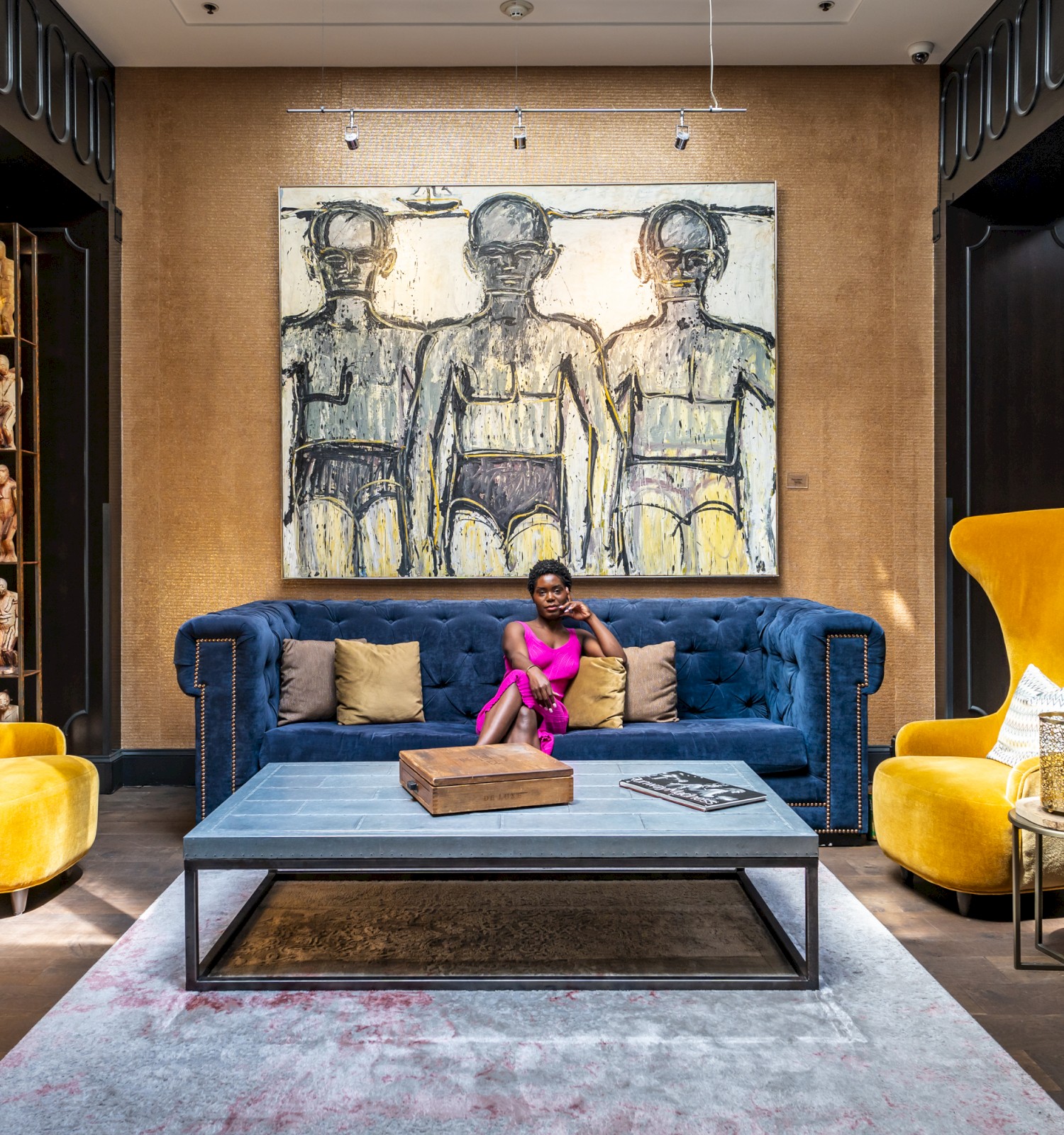 A person sits on a blue couch in a stylish lounge with yellow chairs, a large painting, and elegant decor.