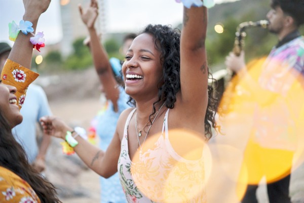 A group of people is joyfully dancing outdoors, with a person playing the saxophone in the background and lights creating a festive atmosphere.