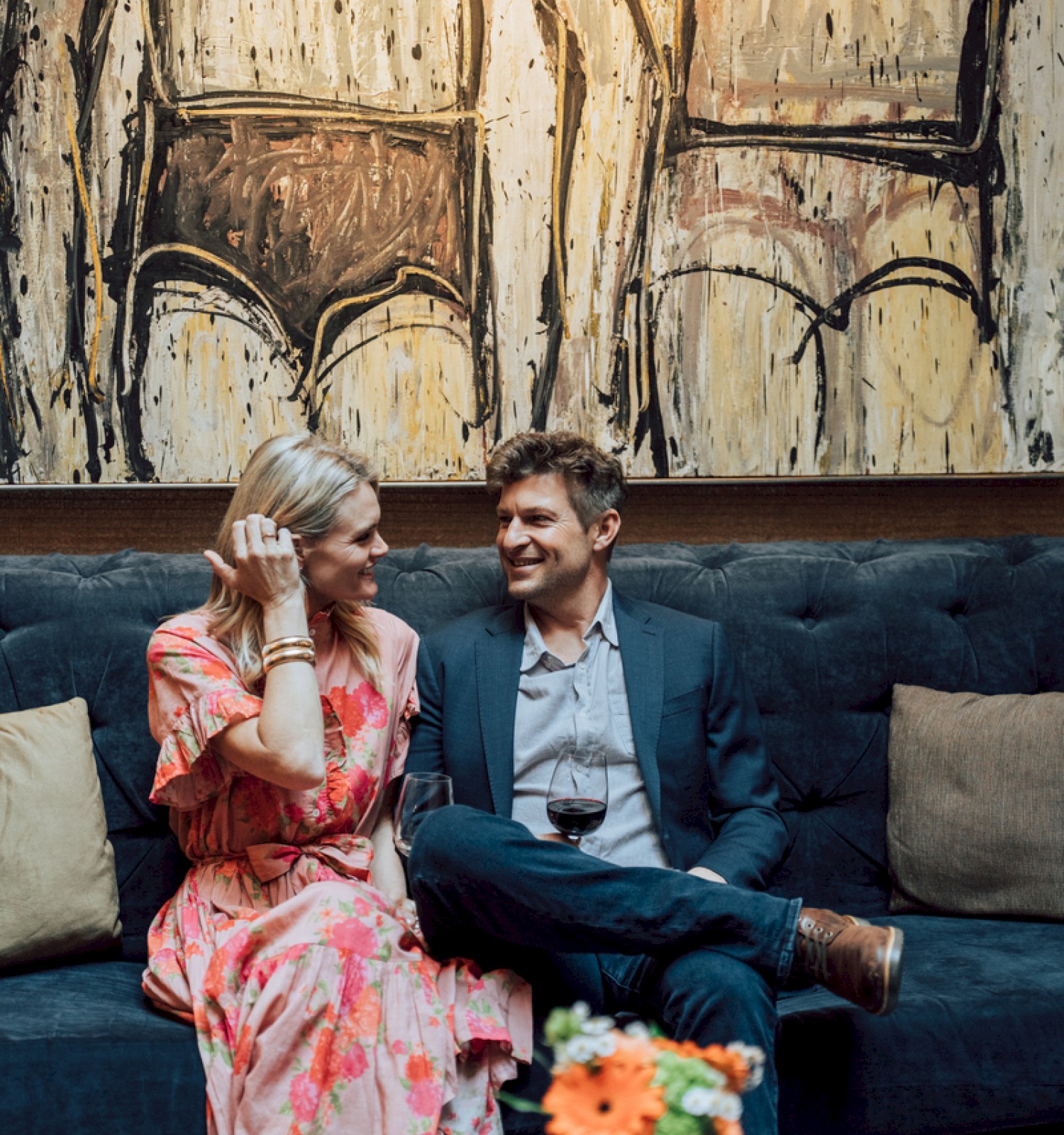 A couple sits on a couch with wine, under abstract art, facing each other and smiling.