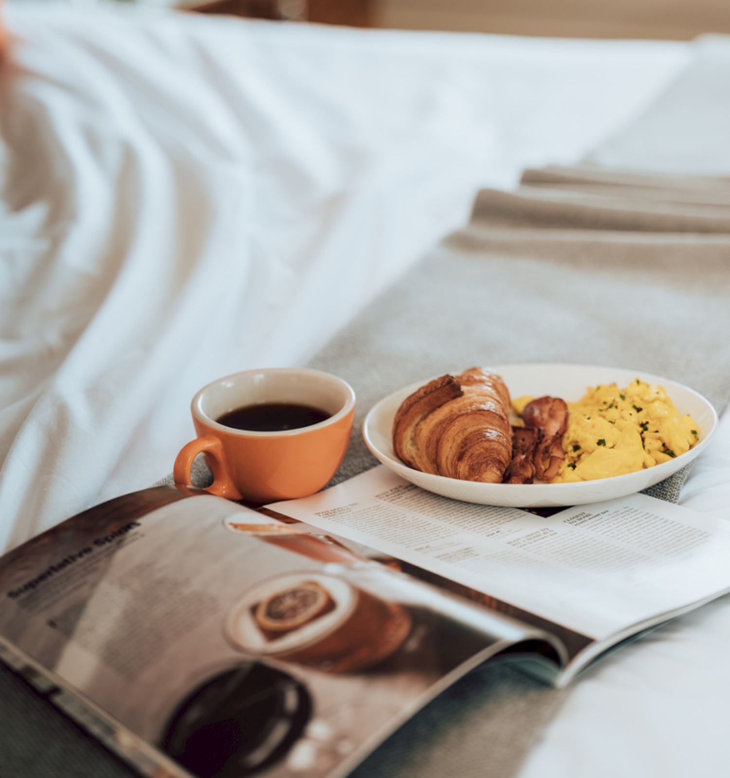 A cozy breakfast scene with coffee, croissant, scrambled eggs, and bacon on a bed, alongside an open magazine.