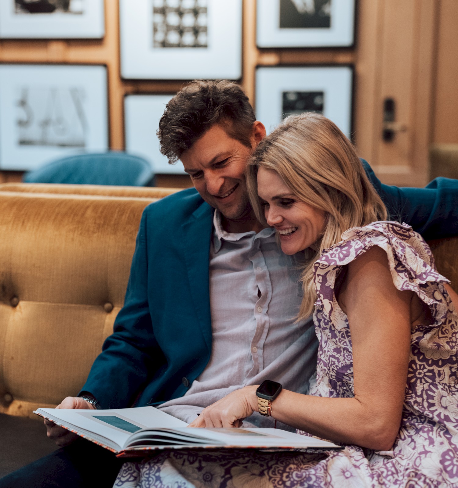 A couple sitting on a couch, looking at a book together, with framed pictures on the wall behind them, creating a cozy atmosphere.