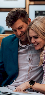 A couple sitting close on a couch, smiling while looking at a book together in a cozy room with framed pictures on the wall.
