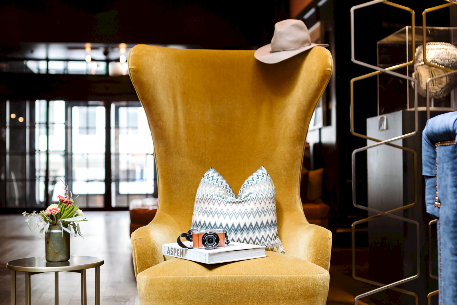A cozy setting with a yellow armchair, hat, pillow, camera on a book, and flowers on a table.