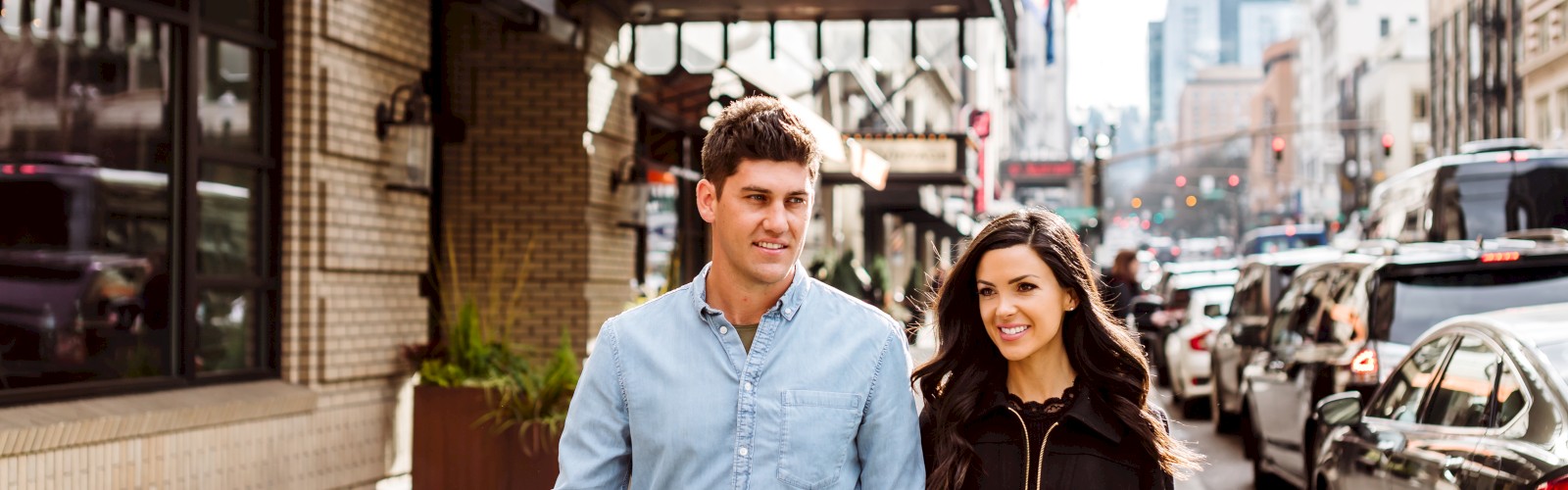 A couple walks down a city street, holding hands, in front of Hotel Lucia, with traffic and buildings in the background.