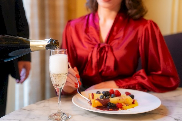 A person in a red blouse enjoys a plate of dessert topped with berries while champagne is being poured into a glass beside them.
