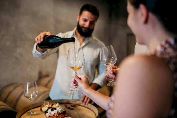 A person is pouring wine into glasses for others in a cellar, with a cheese and fruit platter on a barrel in front of them.