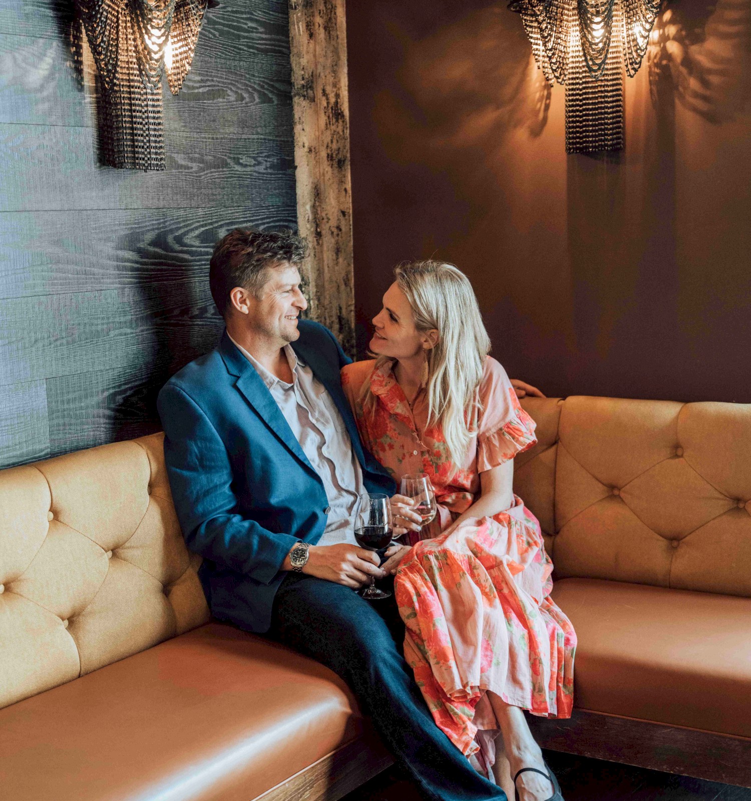 A couple enjoys wine and conversation in a cozy, dimly-lit lounge with decorative wall sconces and warm ambiance.