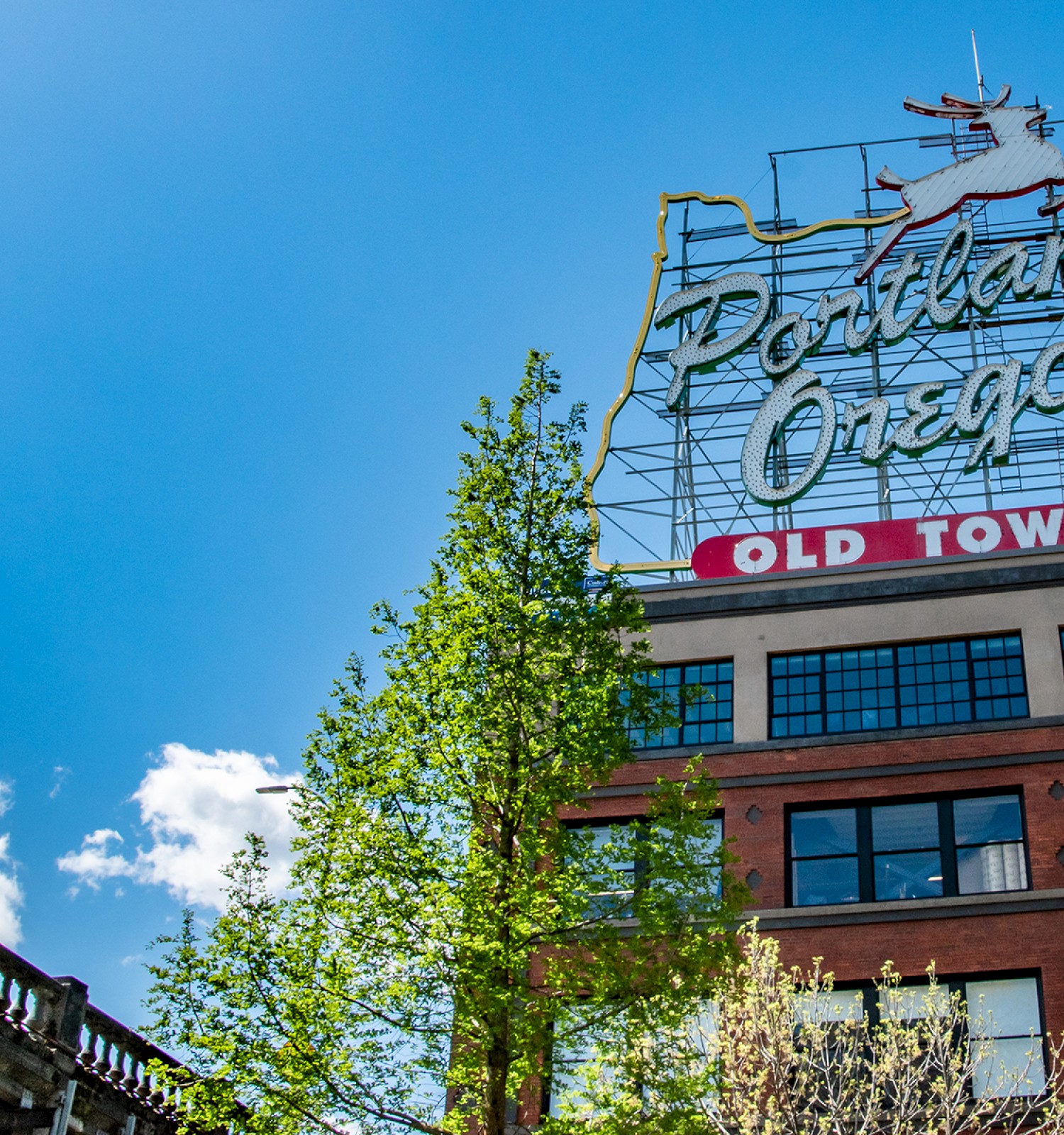 The image shows a sign for Portland, Oregon, with a deer figure on top, all against a bright blue sky with trees and a building below.