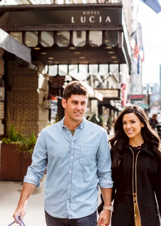 A couple walks hand in hand on a city street near Hotel Lucia, with cars and tall buildings in the background.