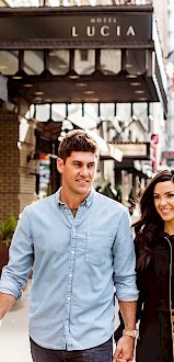 A couple walks down a busy city street, holding hands in the sunshine, with cars parked along the road and buildings visible in the background.
