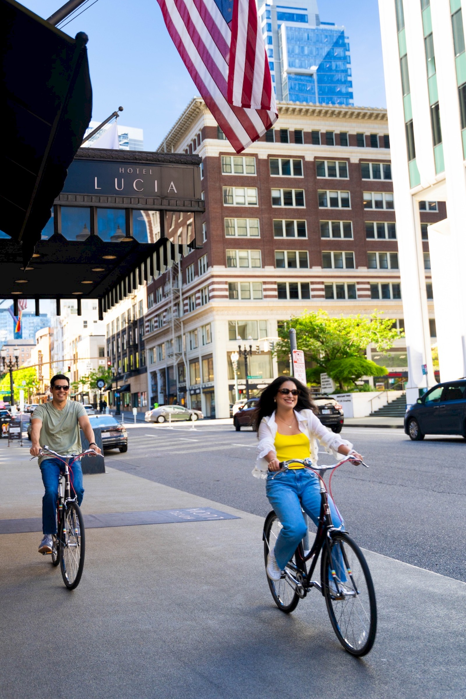 Two people are riding bicycles on a city street with an American flag and tall buildings in the background.