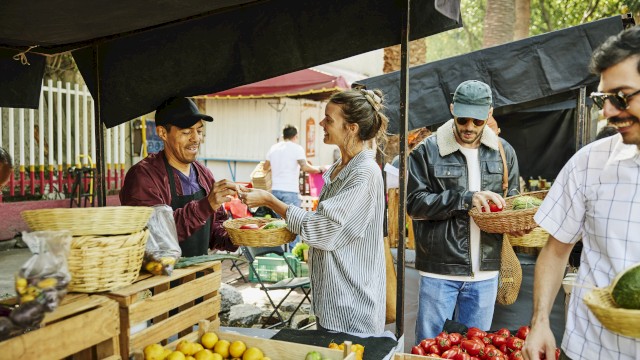 A street market scene with vendors selling citrus and tomatoes, customers browsing baskets under tents, exchanging goods and money.