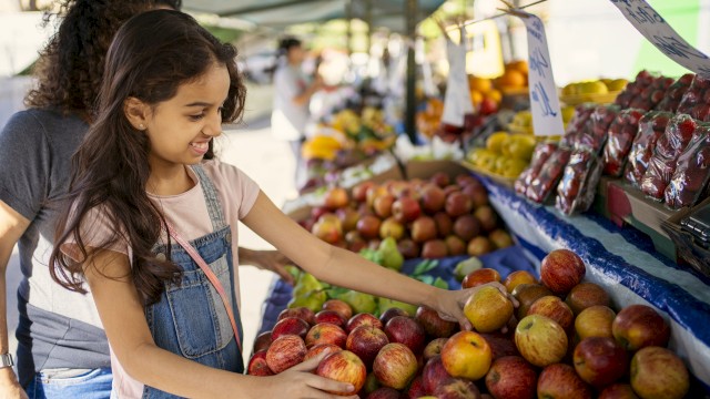 A young girl smiles while selecting apples at a bustling outdoor market stall with fruit and produce around her.