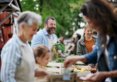 People are at an outdoor market, engaging with fresh produce and smiling in a lively, green setting.