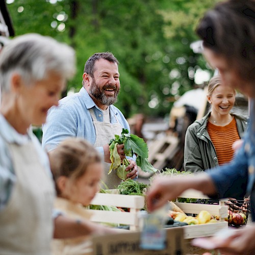 People are at an outdoor market, engaging with fresh produce and smiling in a lively, green setting.