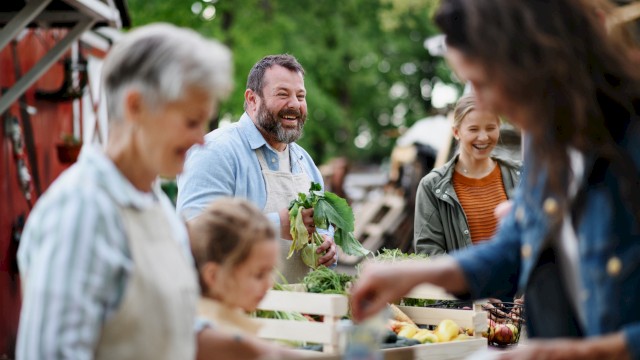 People are at an outdoor market, engaging with fresh produce and smiling in a lively, green setting.