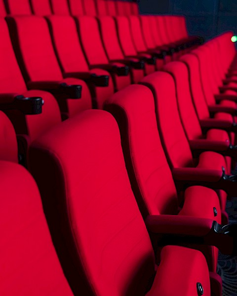 Rows of empty red theater seats in a dark auditorium setting, suggesting a cinema or performance venue.