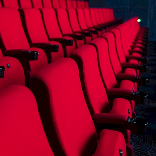 Rows of empty red theater seats in a dark auditorium setting, suggesting a cinema or performance venue.