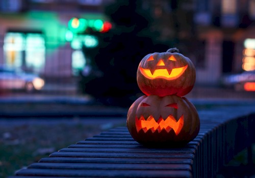 Two lit jack-o’-lanterns stacked on a wooden rail outside at night, with blurred colorful city lights in the background.
