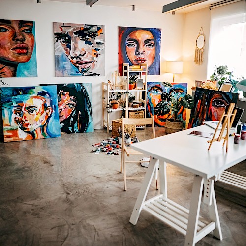 An art studio with vibrant portraits on the walls, a table with painting supplies, and a cozy seating area by the window.