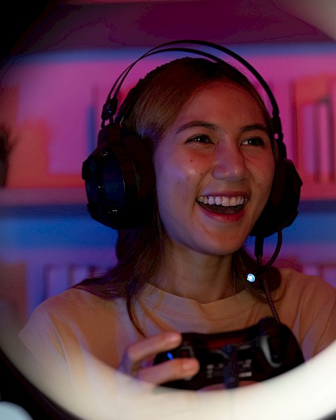 A person is gaming with a headset, smiling and holding a controller, seen through a ring light, in a room with colorful lighting.