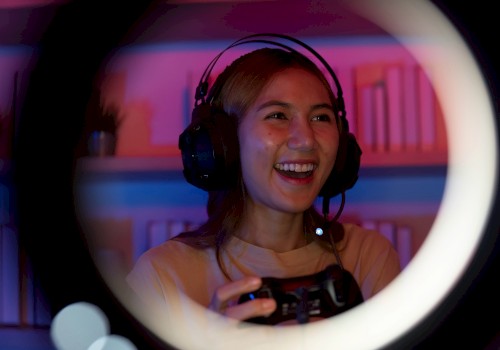 A person is gaming with a headset, smiling and holding a controller, seen through a ring light, in a room with colorful lighting.