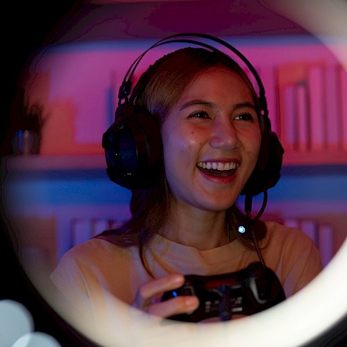 A person is gaming with a headset, smiling and holding a controller, seen through a ring light, in a room with colorful lighting.