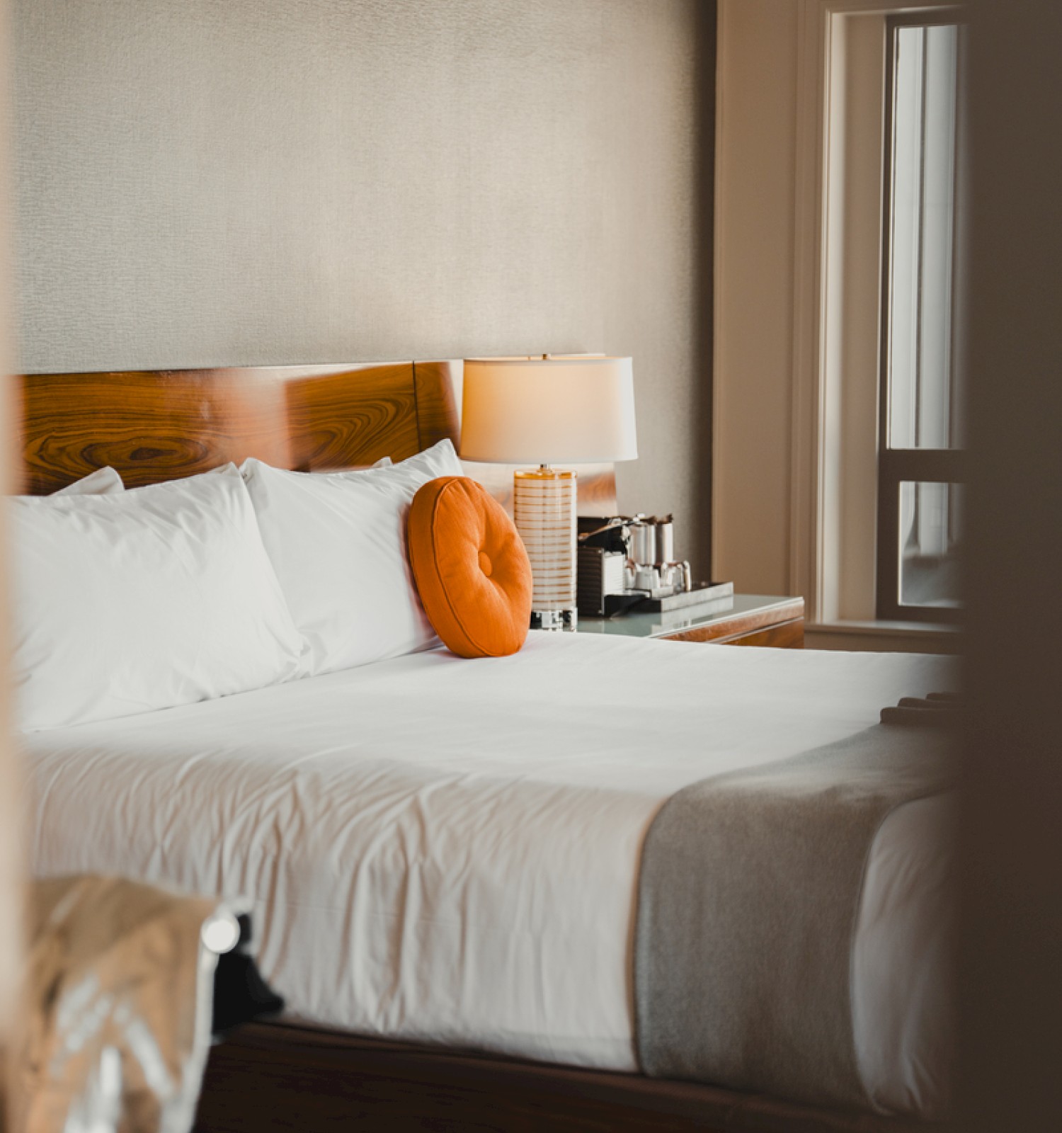 A neatly made bed with white linens, an orange pillow, and a bedside table with a lamp in a cozy room.