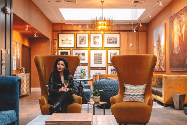A stylish lounge with warm lighting, gallery wall behind, eclectic chairs (two tall orange armchairs), a woman seated, art frames, and comfy decor.