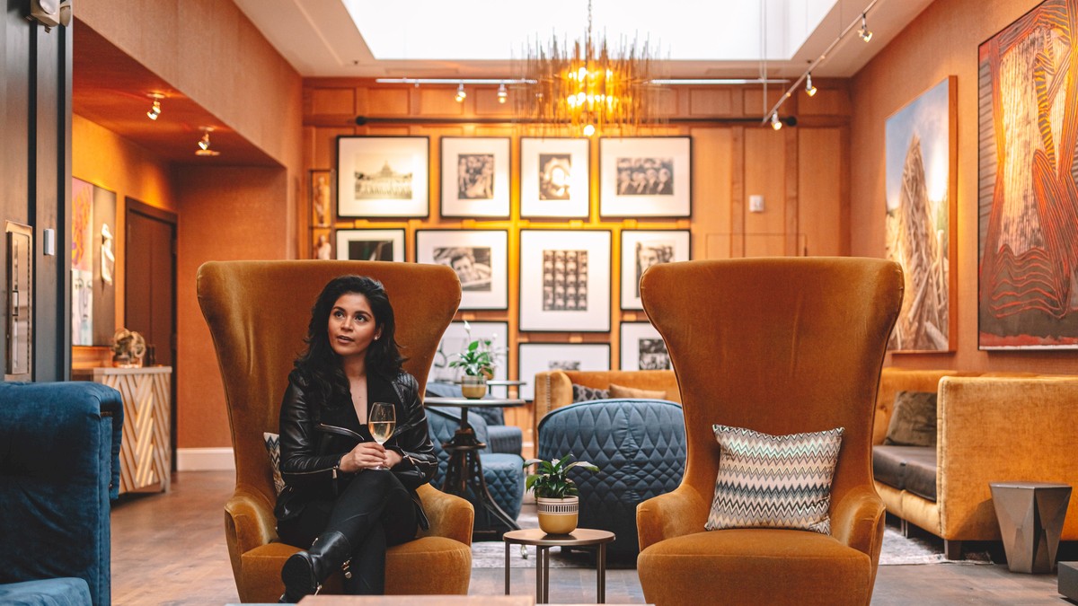 A stylish lounge with warm lighting, art-filled walls, and comfy armchairs; a woman sits holding a mug, surrounded by modern decor.