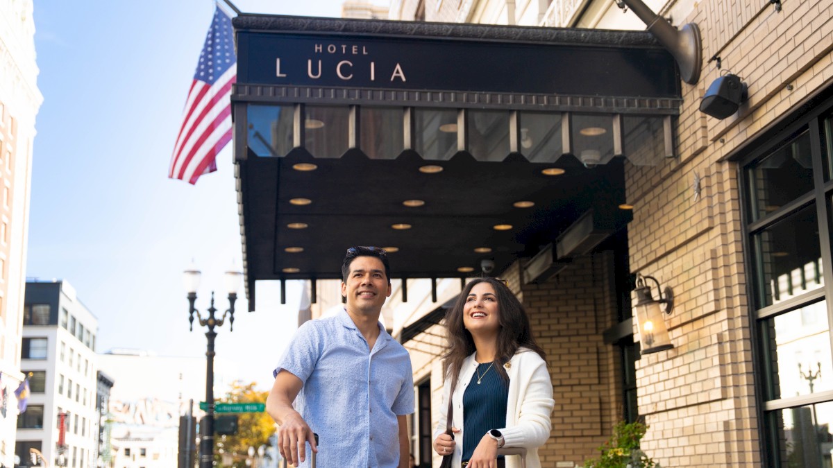 A couple stands smiling under a hotel awning that reads “Hotel Lucia” on a sunny city street, American flag waving nearby.