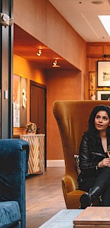 A stylish lobby or lounge with high-backed chairs, art-filled walls, warm lighting, and a woman seated calmly, cigarette in hand, sunglasses.