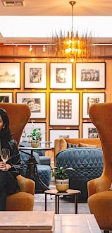 A stylish hotel lobby with warm lighting, two large brown armchairs, a woman sitting center-left, blue and orange seating, framed art on the wall, and a cozy, modern vibe.