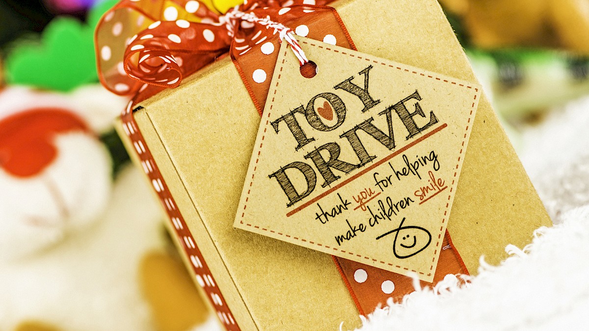 A gift box with a red bow and tag reading “Toy Drive,” teddy bears and colorful holiday decor in the background.