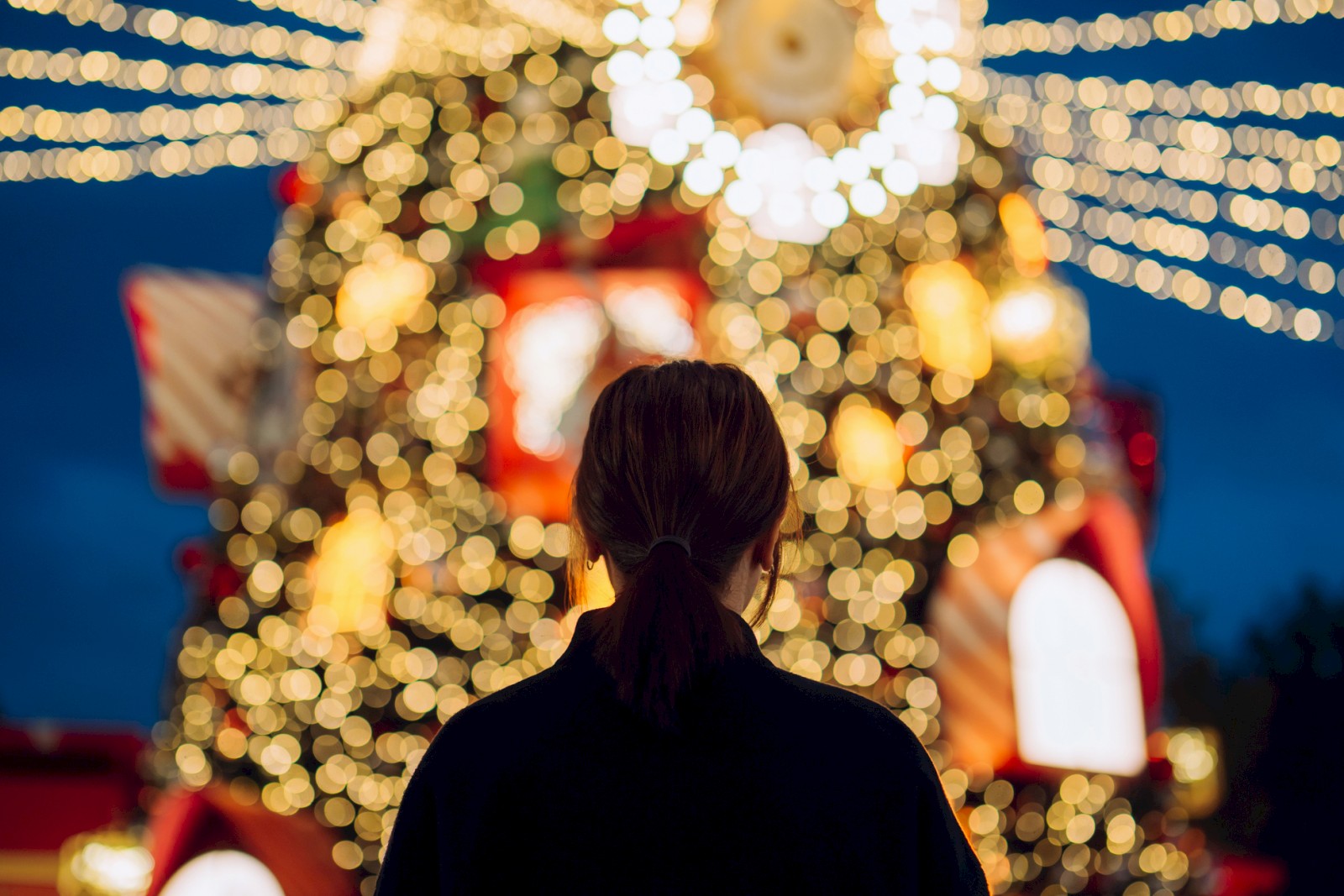 A person stands silhouetted before a large, illuminated Christmas tree with glowing orbs and festive lights in the evening.