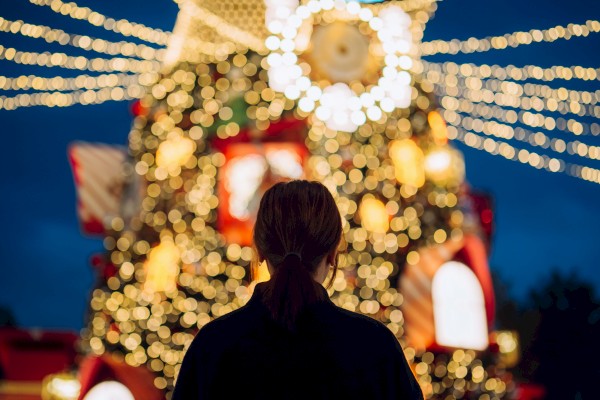 A person stands silhouetted before a large, festive Christmas tree with glowing lights and bokeh in the background.