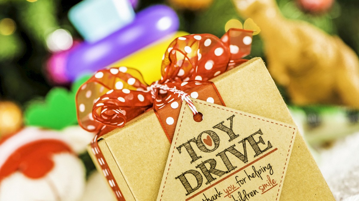 A wrapped gift box with a red ribbon and a “Toy Drive” tag, festive toys blurred in the background, holiday mood, snowy scene.