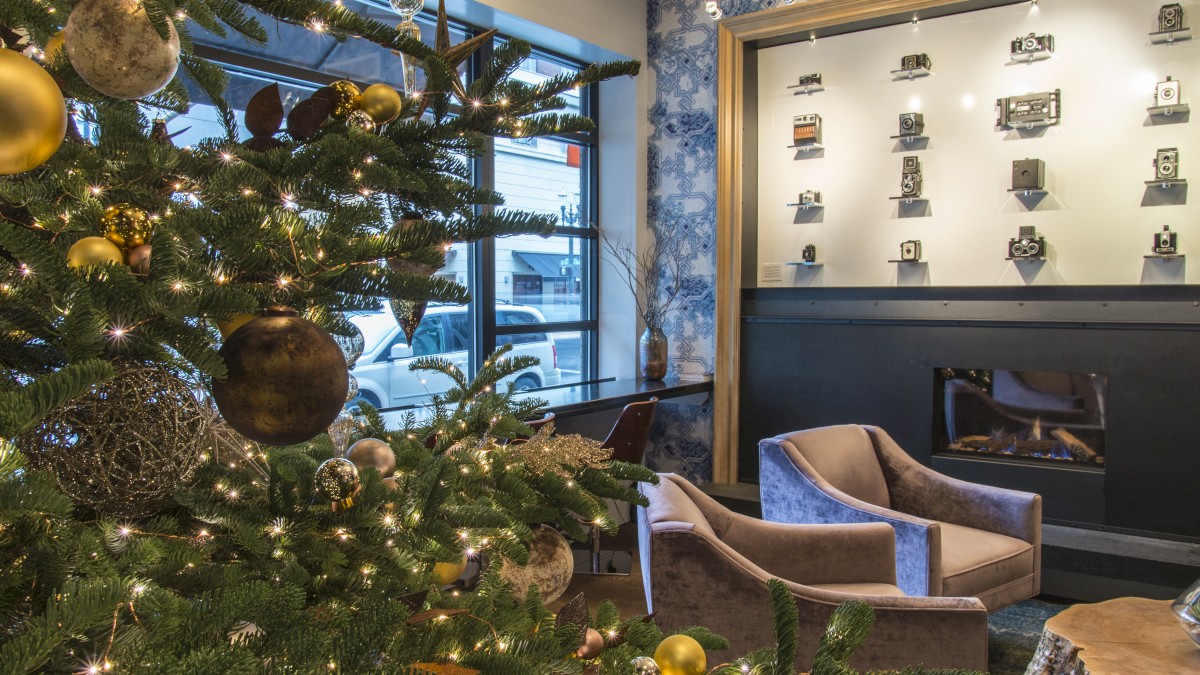A cozy lobby with a decorated Christmas tree, warm seating, and a wall display of small framed items illuminated above a dark booth area.